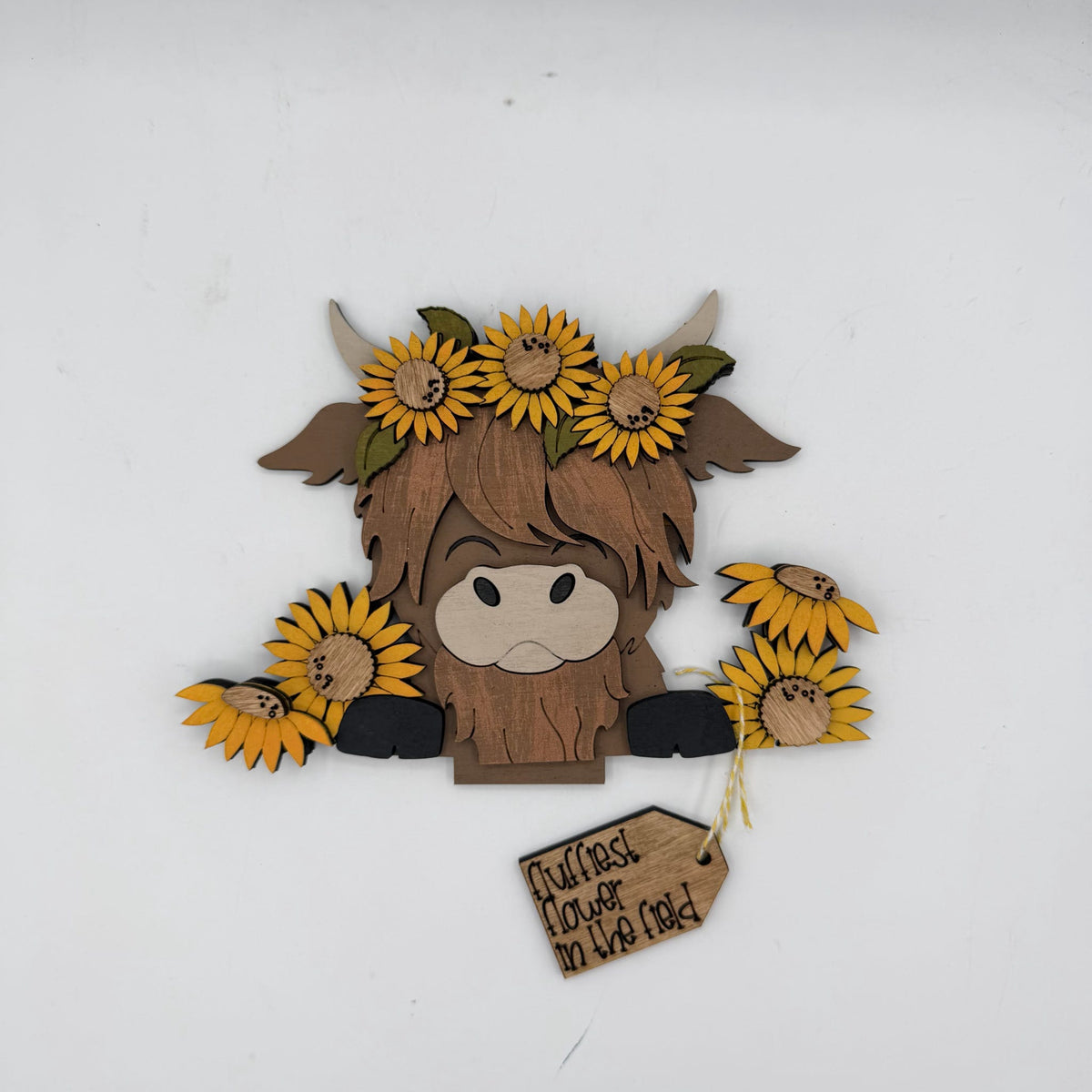 A rustic wooden Highland cow decoration with yellow sunflower accents, a light wood face, and a hanging tag that reads 'Rustic Flower in the Mud' on a plain white background.