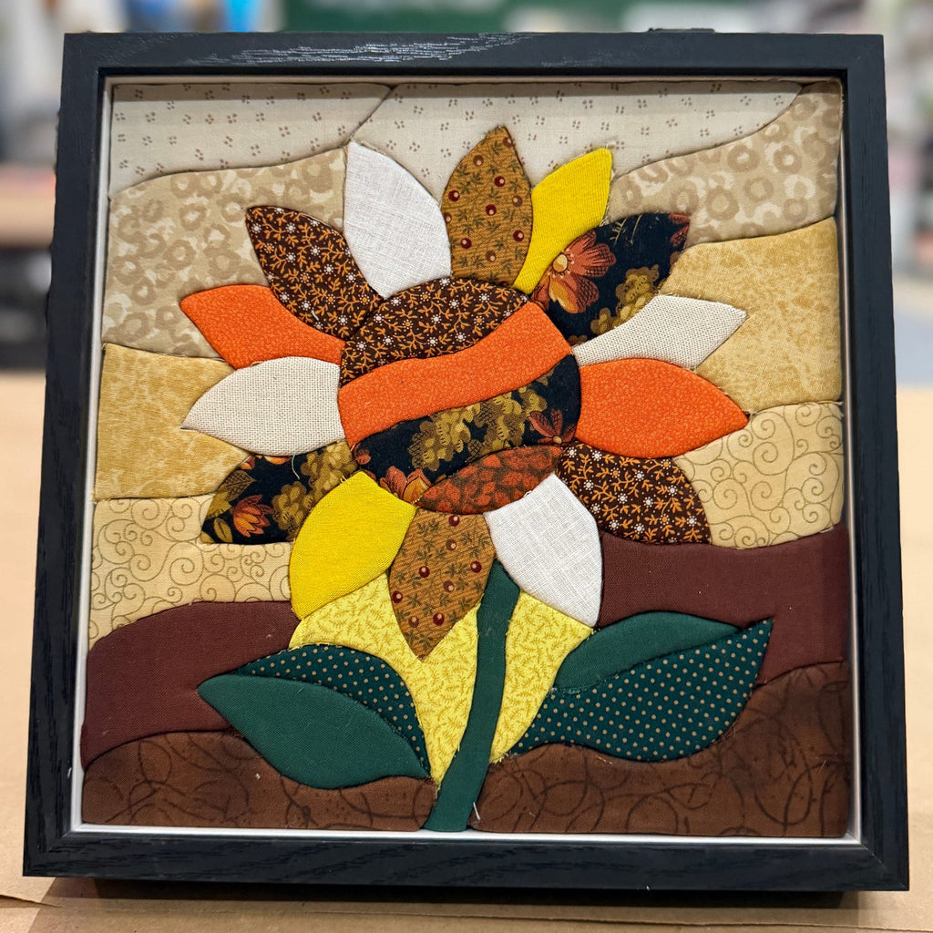 A framed quilted fabric sunflower artwork with layered petals in orange, yellow, white, and brown, set in a black frame on a tabletop with a blurred indoor background.