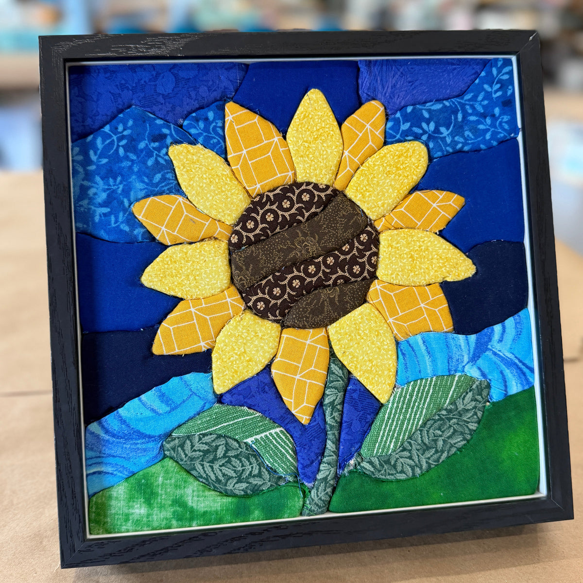 A framed fabric art piece featuring a stylized sunflower with yellow petals, a brown patterned center, blue and dark blue background shapes, and green leaves on a tan surface.