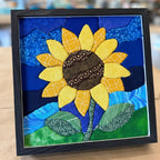 A framed fabric art piece featuring a stylized sunflower with yellow petals, a brown patterned center, blue and dark blue background shapes, and green leaves on a tan surface.