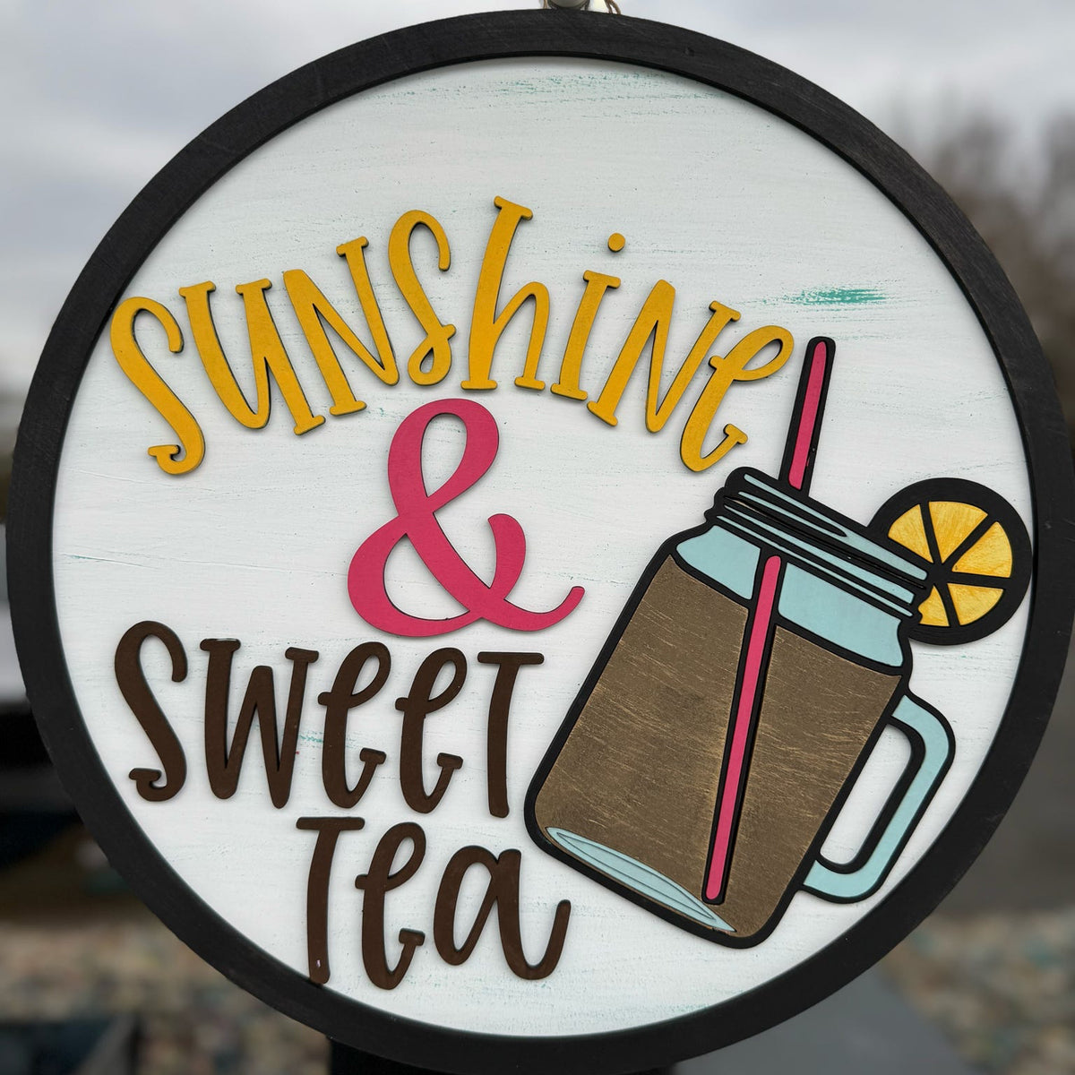 A door hanger with a circular shape featuring a graphic design with text 'Sunshine & Sweet Tea' and an illustration of a tea cup with a lemon slice and a straw.
