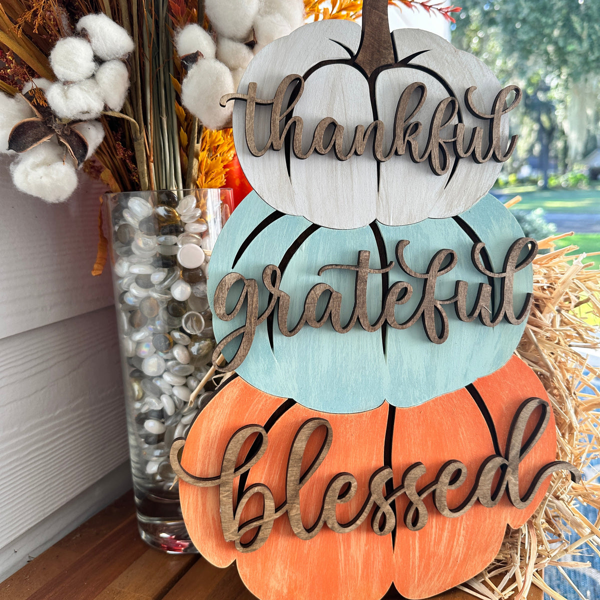A decorative trio of stacked pumpkins with the words 'Thankful', 'Grateful', and 'Blessed' carved into their surfaces, placed on a wooden surface with autumn-themed decorations around.