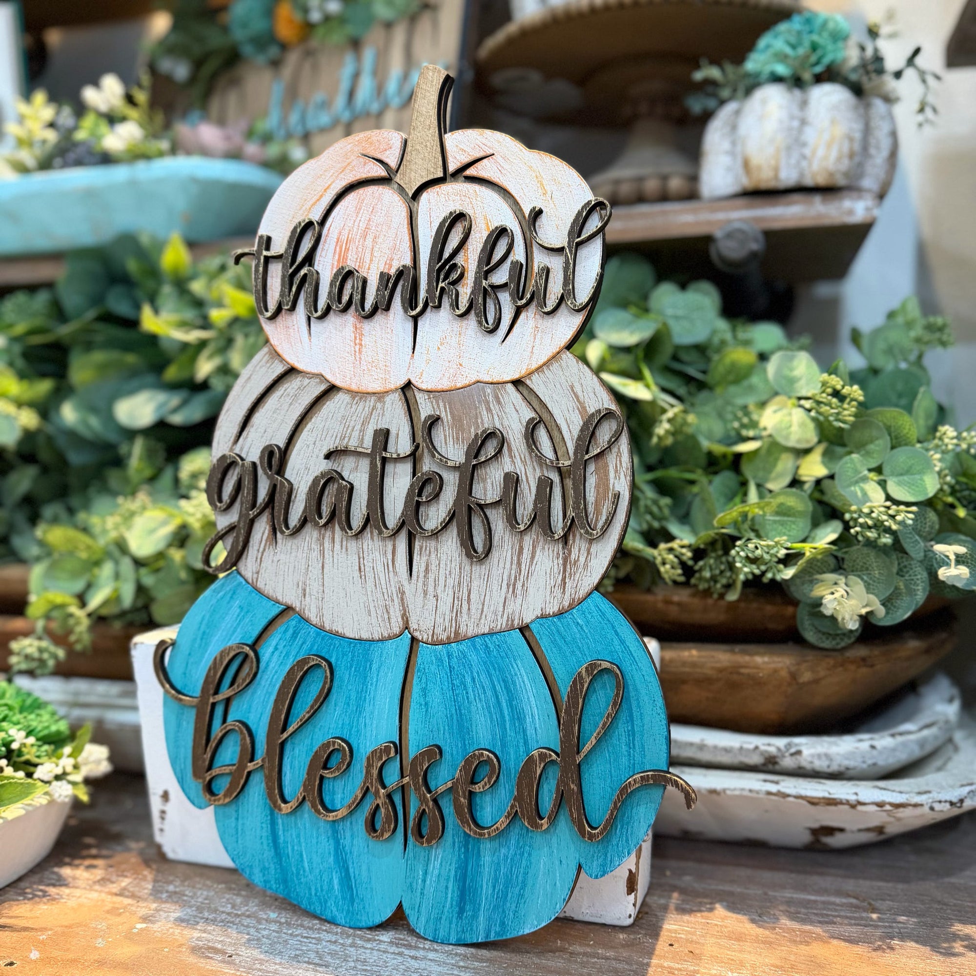 A decorative stacked autumn sign shaped like three pumpkins with the words 'thankful', 'grateful', and 'blessed' in cursive script. The top pumpkin is off-white, the middle is weathered wood-toned, and the bottom is bright turquoise.