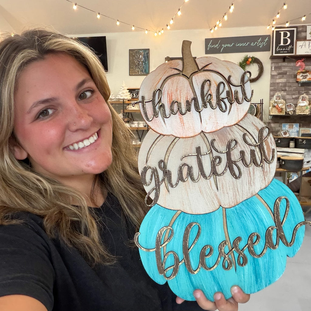 A smiling woman holds a decorative autumn sign shaped like three stacked pumpkins. The sign has the words 'thankful', 'grateful', and 'blessed' in dark script over turquoise, cream, and light peach pumpkins.