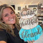 A smiling woman holds a decorative autumn sign shaped like three stacked pumpkins. The sign has the words 'thankful', 'grateful', and 'blessed' in dark script over turquoise, cream, and light peach pumpkins.