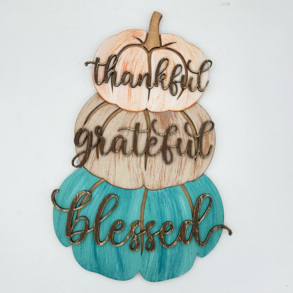 A decorative autumn wall sign featuring three stacked pumpkins in peach, tan, and teal, with the words 'thankful', 'grateful', and 'blessed' cut out in brown script across the front, shown on a plain white background.