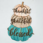 A decorative autumn wall sign featuring three stacked pumpkins in peach, tan, and teal, with the words 'thankful', 'grateful', and 'blessed' cut out in brown script across the front, shown on a plain white background.