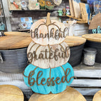 A decorative stacked pumpkin sign with the words 'thankful', 'grateful', and 'blessed' in raised script, displayed on a rustic wooden shelf in a craft or home decor store. The top two pumpkins are natural wood, and the bottom pumpkin is painted turquoise blue.