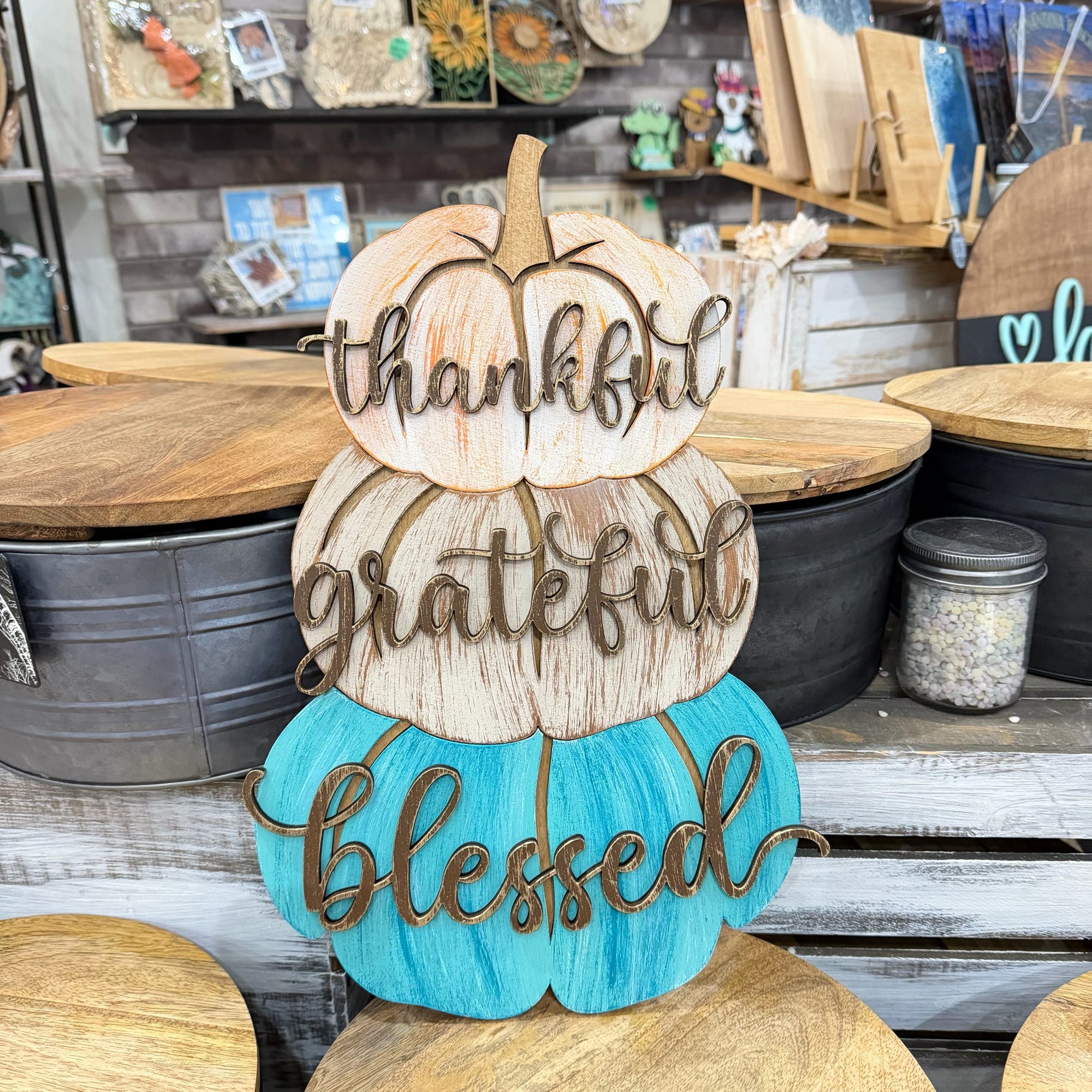 A decorative stacked pumpkin sign with the words 'thankful', 'grateful', and 'blessed' in raised script, displayed on a rustic wooden shelf in a craft or home decor store. The top two pumpkins are natural wood, and the bottom pumpkin is painted turquoise blue.