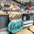 A wooden decorative sign shaped like stacked pumpkins with the words 'thankful', 'grateful', and 'blessed' in raised script, displayed in a rustic shop setting.