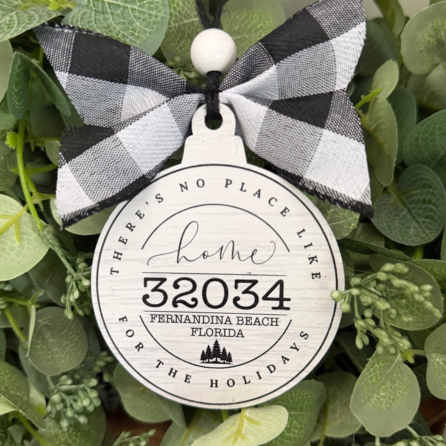 A round white holiday ornament with black text reading 'home 32034 Fernandina Beach Florida' and 'There is no place like for the holidays,' hanging from a black ribbon bow with a black-and-white buffalo plaid pattern and a white bead, displayed among green eucalyptus leaves.