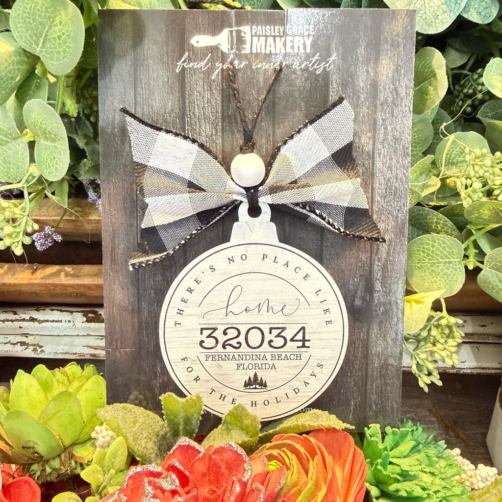 A rustic wooden hanging ornament card from Paisley Creek Makersy, featuring a buffalo plaid bow and a round tag that reads 'Home 32034 Fernandina Beach Florida' surrounded by decorative succulents and flowers.