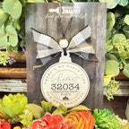 A rustic wooden hanging ornament card from Paisley Creek Makersy, featuring a buffalo plaid bow and a round tag that reads 'Home 32034 Fernandina Beach Florida' surrounded by decorative succulents and flowers.