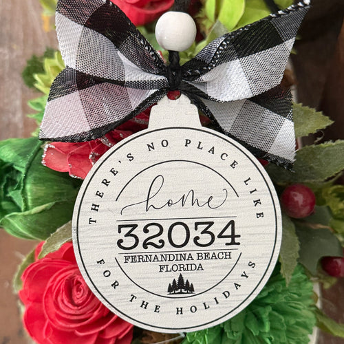A round white holiday ornament with black text reading 'Home 32034 Fernandina Beach Florida' and 'There's no place like for the holidays,' tied with a black-and-white gingham ribbon bow and surrounded by red flowers and green leaves.