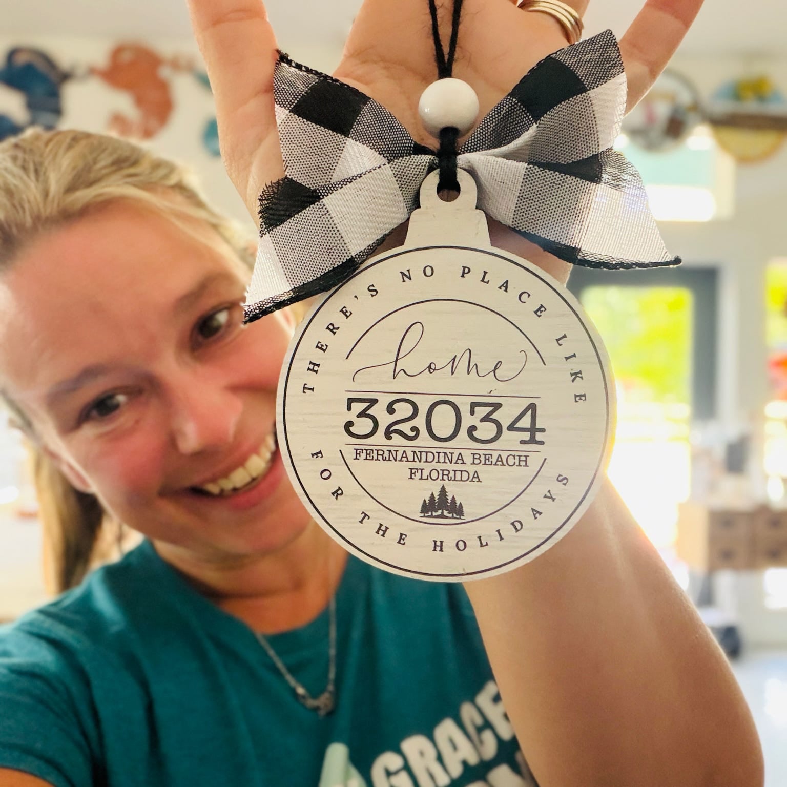 A woman holds up a personalized Christmas ornament featuring a black and white buffalo plaid bow and a round white tag with custom text reading 'Home 32034 Fernandina Beach Florida' on a blurred indoor background.