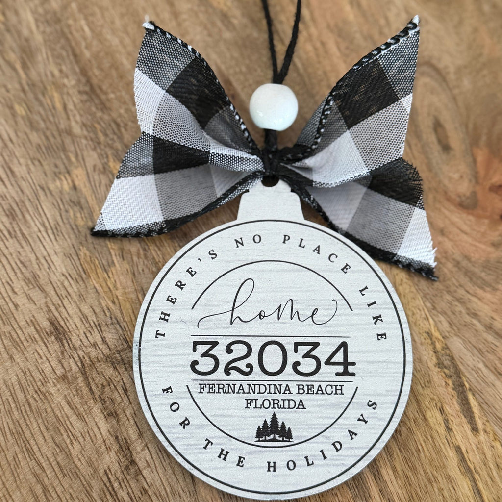 A personalized holiday ornament featuring a black and white buffalo plaid ribbon bow and a round white tag printed with 'Home 32034 Fernandina Beach Florida' on a rustic wooden background.