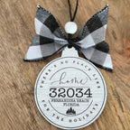A personalized holiday ornament featuring a black and white buffalo plaid ribbon bow and a round white tag printed with 'Home 32034 Fernandina Beach Florida' on a rustic wooden background.