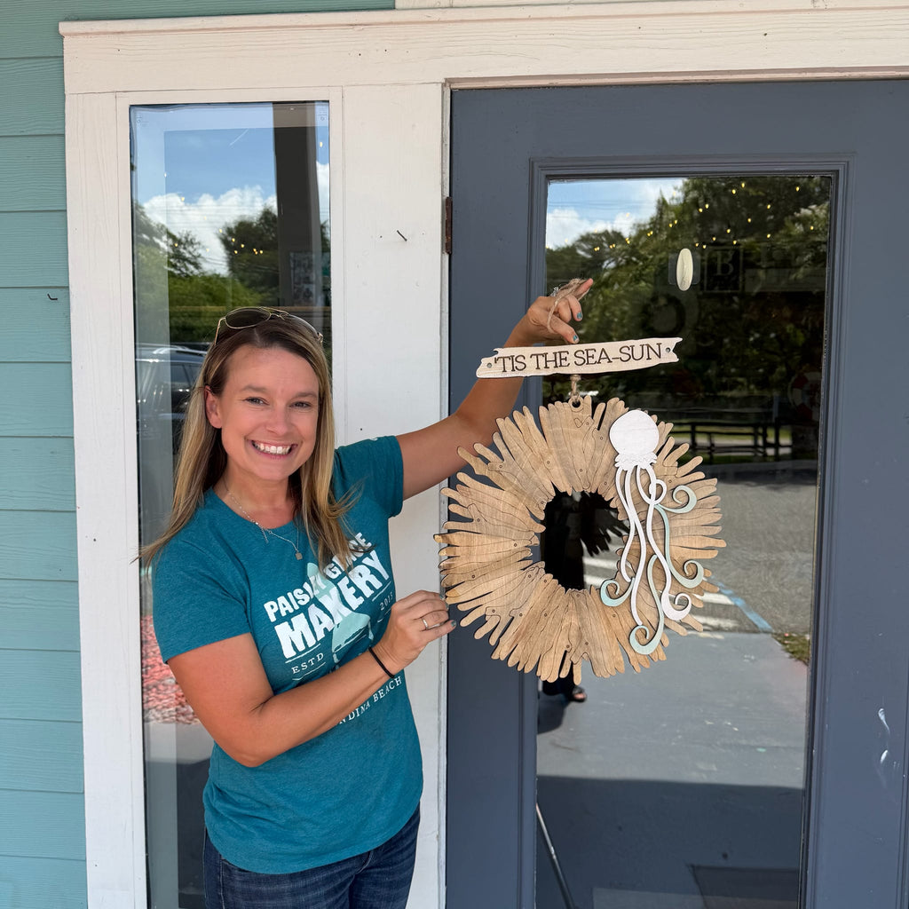A woman smiling and holding a decorative nautical wreath in front of a gray door. The wreath is made from driftwood and has a white cutout of the Sea God triquetra and a wooden sign that reads 'IT'S THE SEA-SUN'.