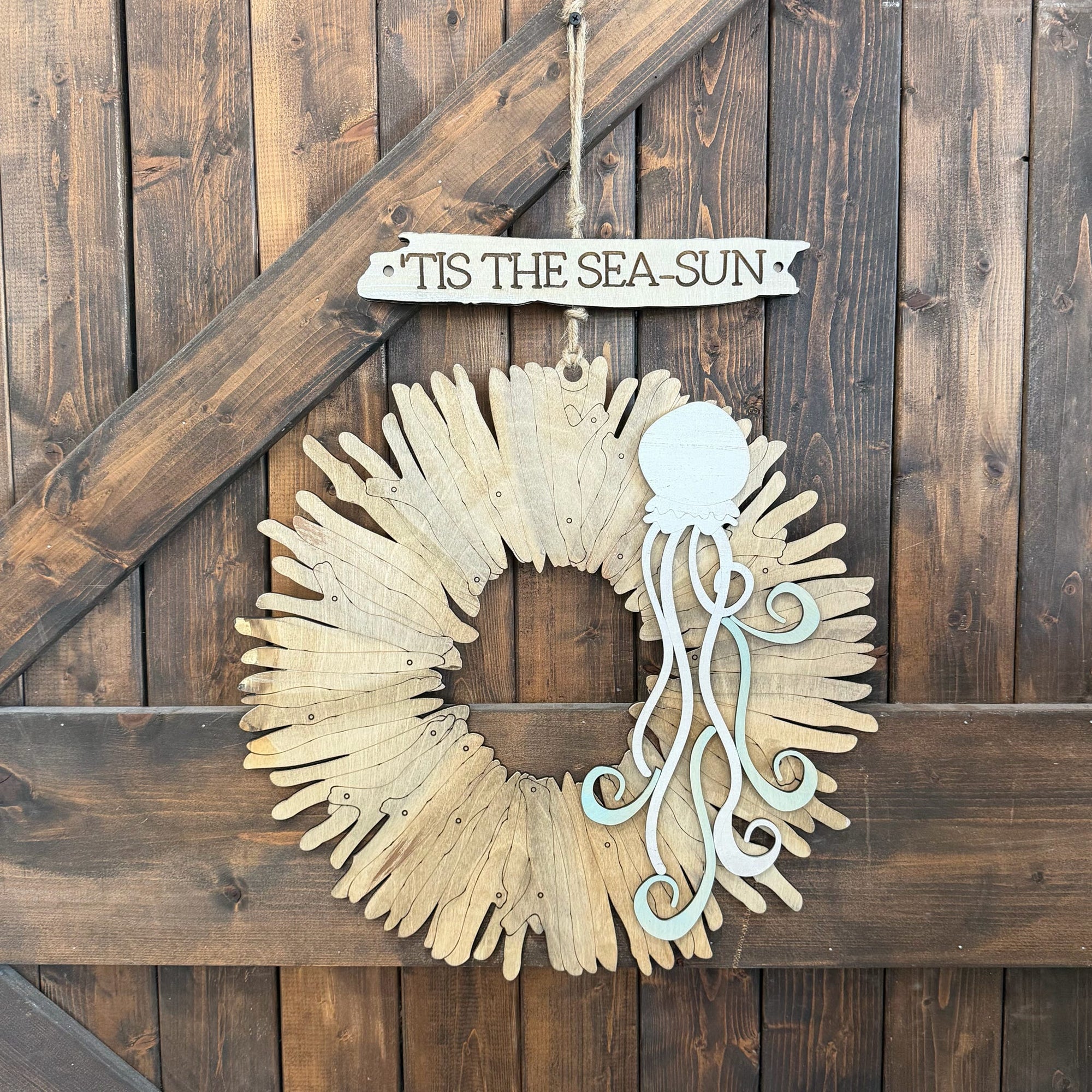 A rustic nautical wall hanging featuring a circular wreath made from light wood driftwood pieces, with a pale blue octopus decoration and a hanging sign that reads “TIS THE SEA-SUN.” The wreath is displayed against a dark brown wooden plank wall.