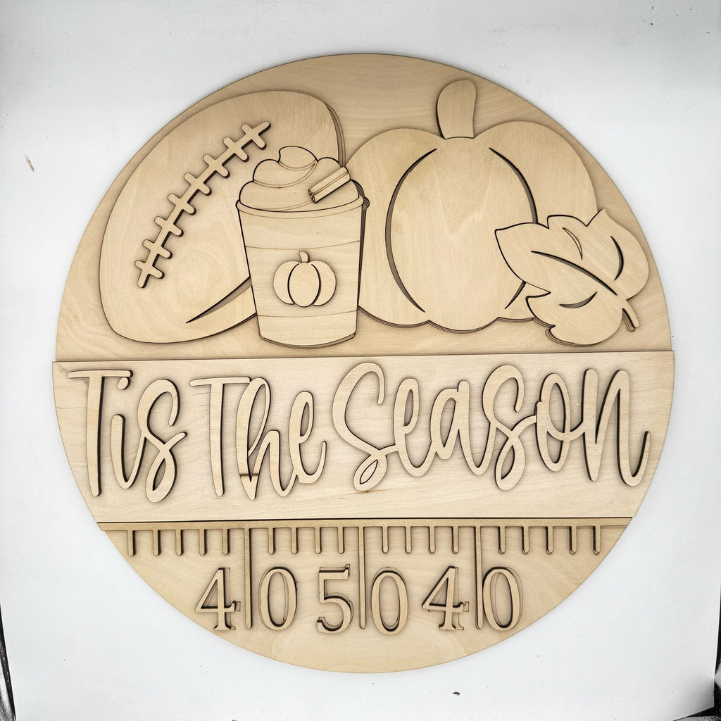 A round wooden plaque featuring engraved autumn-themed illustrations including a football, pumpkin, hot cocoa cup, leaf, and the phrase 'This is the Season' above a measurement scale. The plaque is photographed on a plain white background.