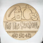 A round wooden plaque featuring engraved autumn-themed illustrations including a football, pumpkin, hot cocoa cup, leaf, and the phrase 'This is the Season' above a measurement scale. The plaque is photographed on a plain white background.
