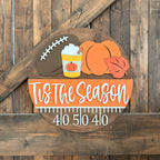 A rustic autumn-themed wooden yard sign hanging on a dark wood plank wall. The sign features a football, a pumpkin, a leaf, a drink with whipped cream, and the text 'Tis The Season' above a football yard line measurement with the numbers '40 50 40'.