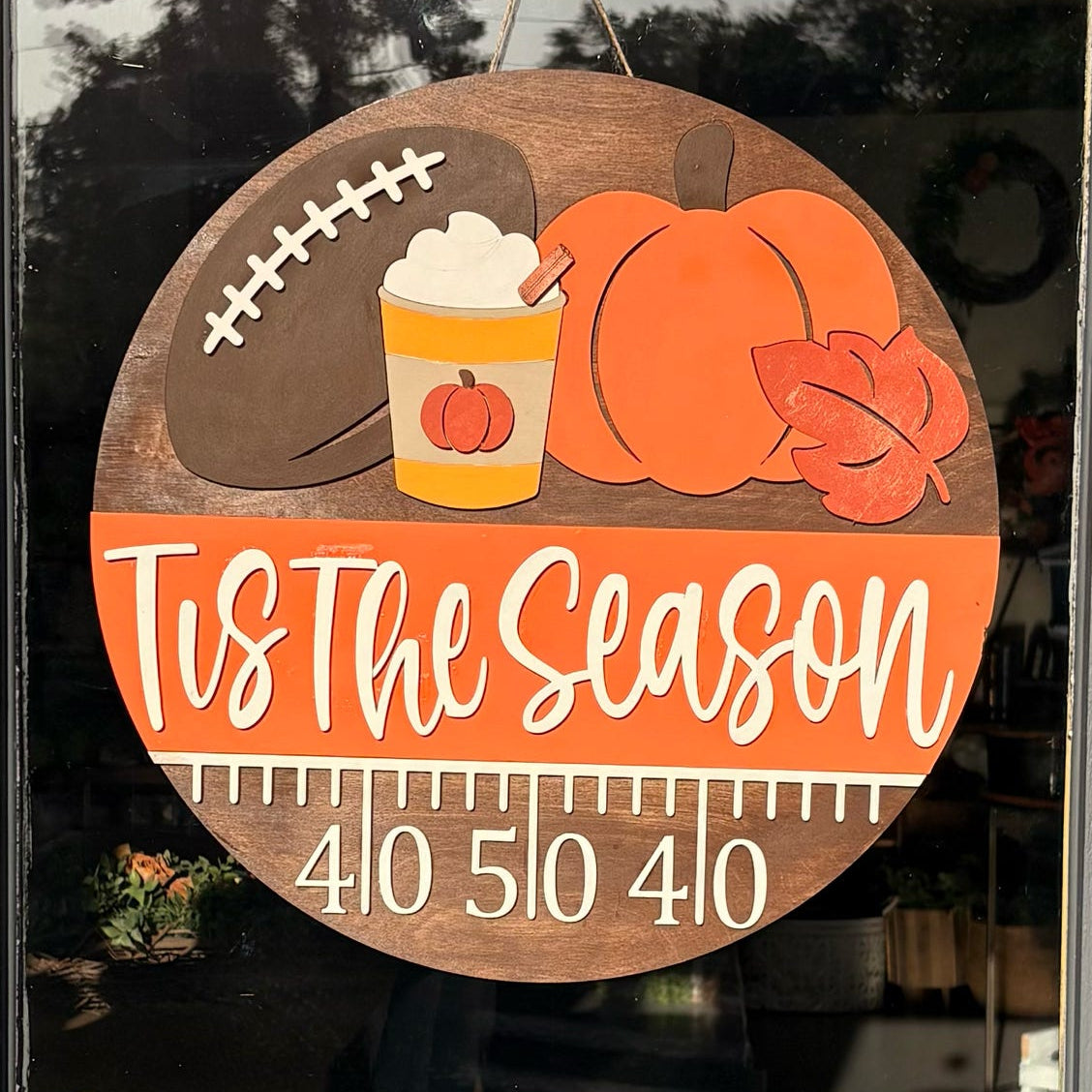 A round wooden autumn-themed door sign hanging on a glass door. The sign features illustrations of a football, pumpkins, a hot chocolate cup, and a leaf, with the text 'Tis The Season' and a measurement scale reading 410, 510, 410.