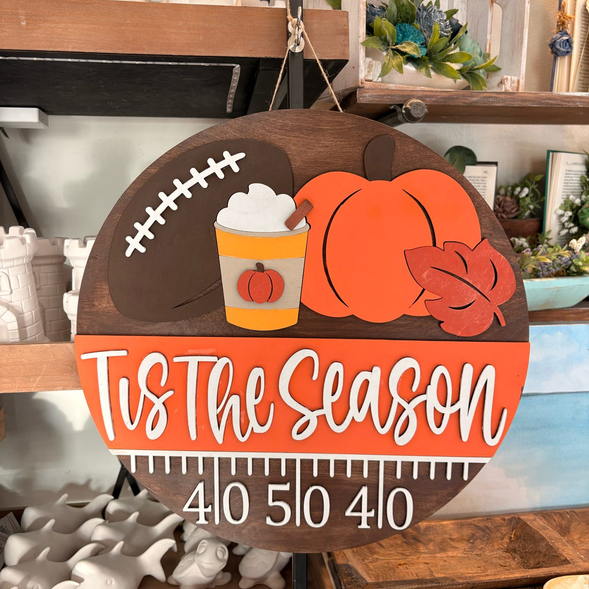 A round wooden seasonal wall sign hanging in a store. The sign is dark brown wood with a printed autumn football-themed design featuring orange pumpkins, a brown football, a yellow drink with whipped cream, and a leaf.