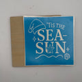 A square blue sticker or decal with white text reading "'Tis the Sea-Sun" and sea-themed illustrations, shown on a tan backing sheet against a light gray background.