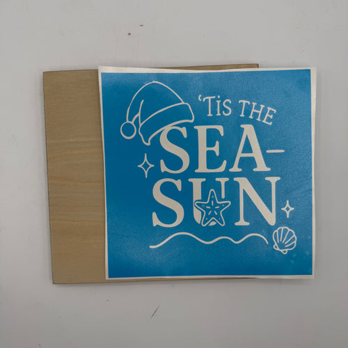 A square blue sticker or decal with white text reading "'Tis the Sea-Sun" and sea-themed illustrations, shown on a tan backing sheet against a light gray background.