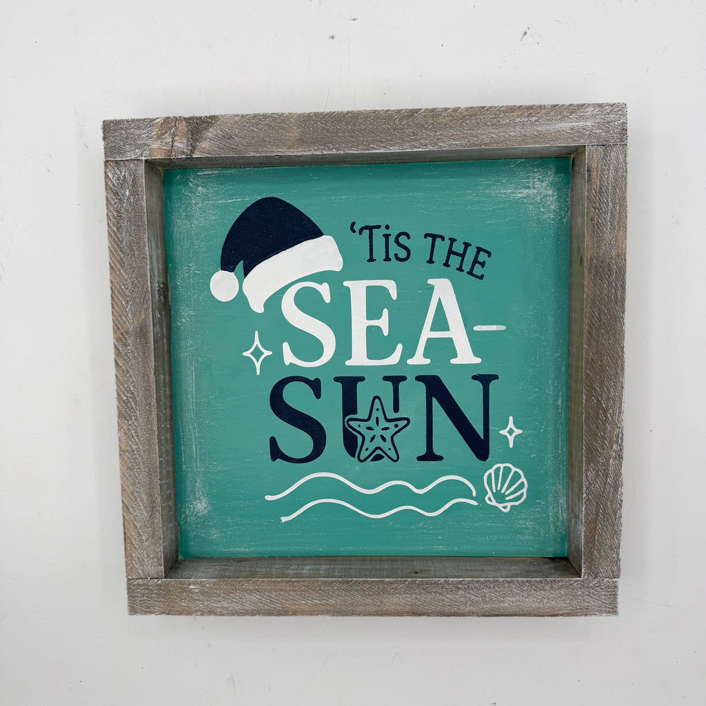 A square framed rustic wall sign with a weathered gray-brown wooden frame and a teal painted center. The sign reads "'Tis the Sea Sun" in white and dark navy lettering, decorated with a navy Santa hat, white seashells and wavy lines, and a white starfish.