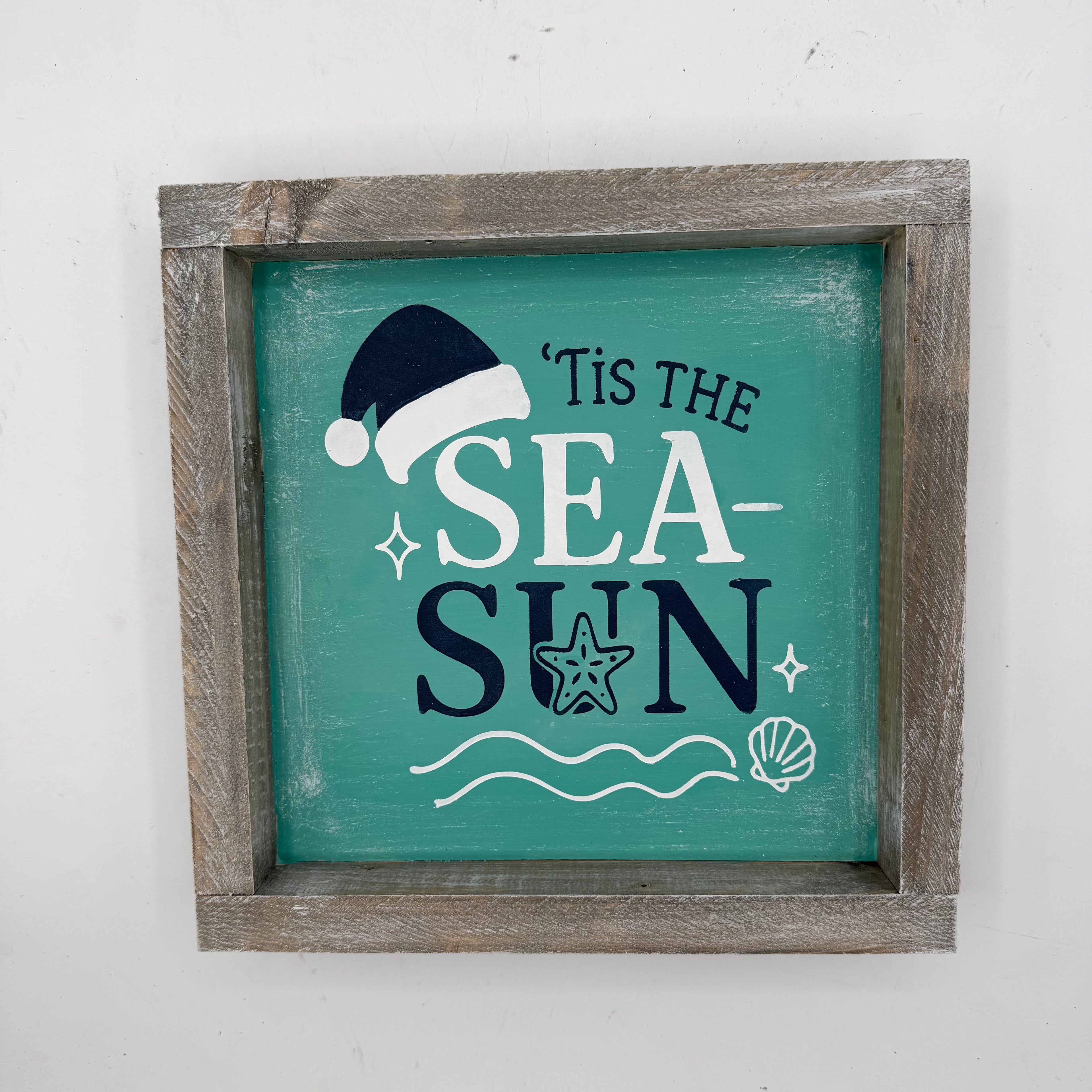 A square framed rustic wall sign with a weathered gray-brown wooden frame and a teal painted center. The sign reads "'Tis the Sea Sun" in white and dark navy lettering, decorated with a navy Santa hat, white seashells and wavy lines, and a white starfish.
