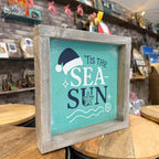 A square wooden framed sign with a distressed gray wood frame and a turquoise background, featuring the text "Tis the Sea Sun" with a Santa hat, starfish, and shell illustrations. The sign is displayed on a wooden table in a shop setting.