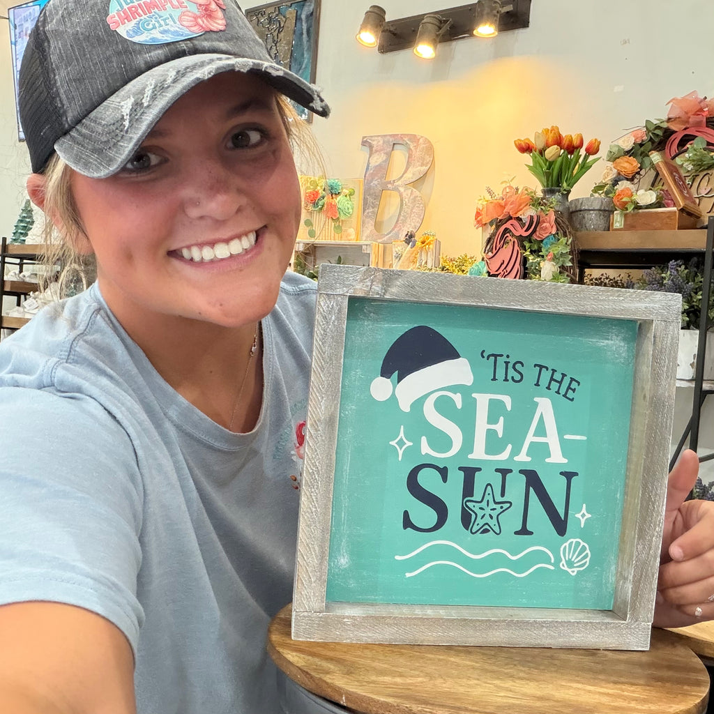 A woman in a gray trucker hat and light blue t-shirt holds up a framed decorative sign that reads 'Tis the Sea Sun' with a Christmas Santa hat and starfish design. The background shows a rustic shop interior with flowers and wooden decor.