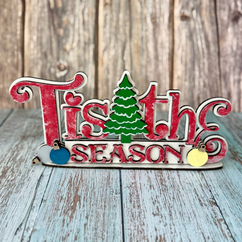 Decorative sign with 'Tis the Season' text and Christmas tree on a wooden background