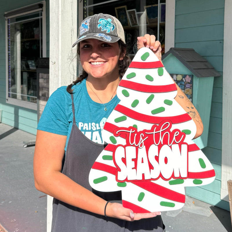 Person holding a decorative Christmas tree sign in front of a building