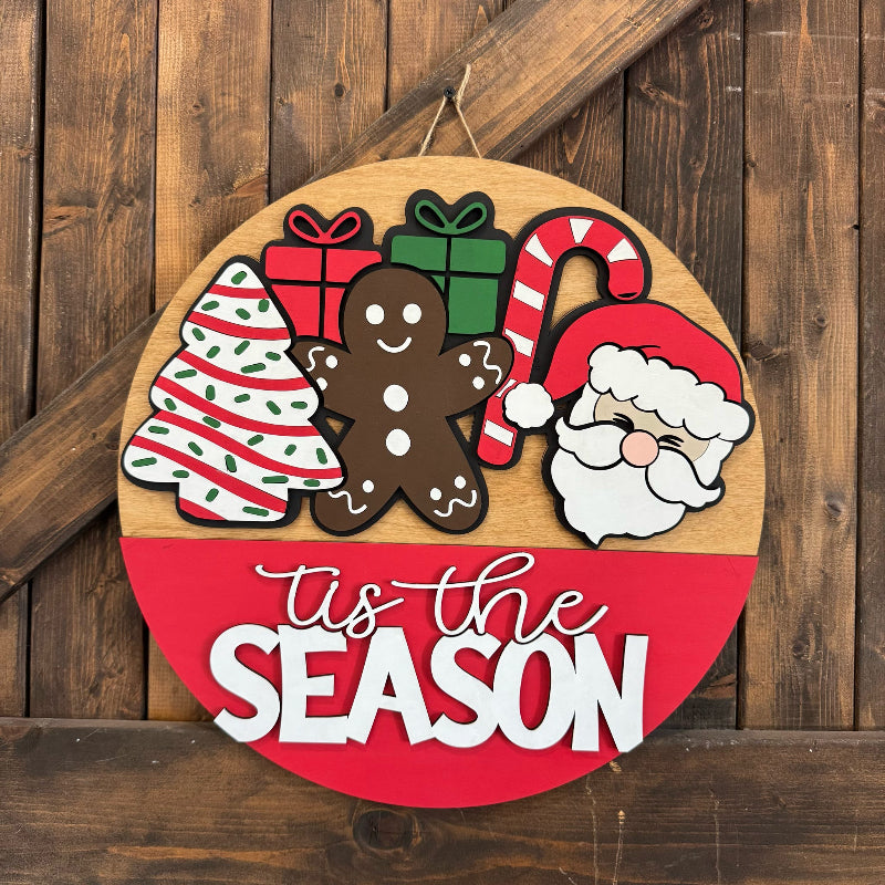 Round wooden sign with Christmas-themed decorations and 'tis the season text on a wooden background.