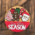 Round wooden sign with Christmas-themed decorations and 'tis the season text on a wooden background.