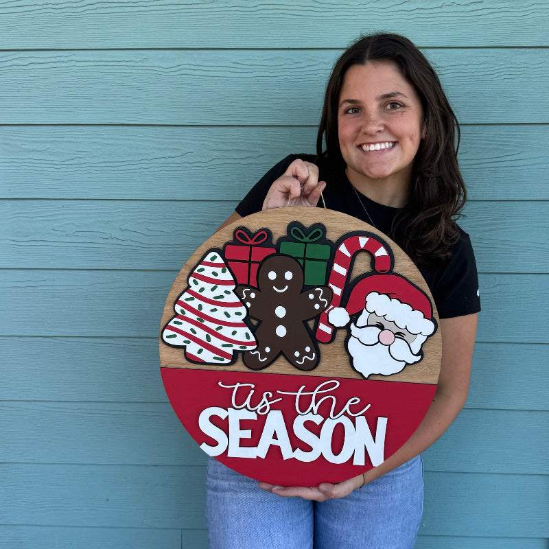 Person holding a Christmas-themed wooden sign with decorative elements and text against a blue wall.