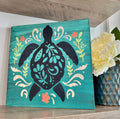 A mini turtle folk art design displayed on a shelf, featuring a silhouette of a turtle with floral patterns in black, green, and orange colors.