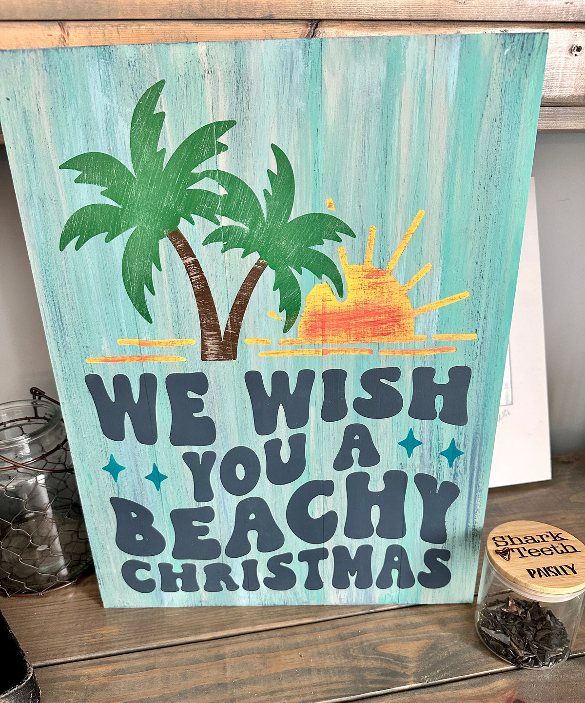 A decorative sign with palm trees and a sun, with text 'WE WISH YOU A BEACHY Christmas', against a turquoise background, displayed on a wooden surface.
