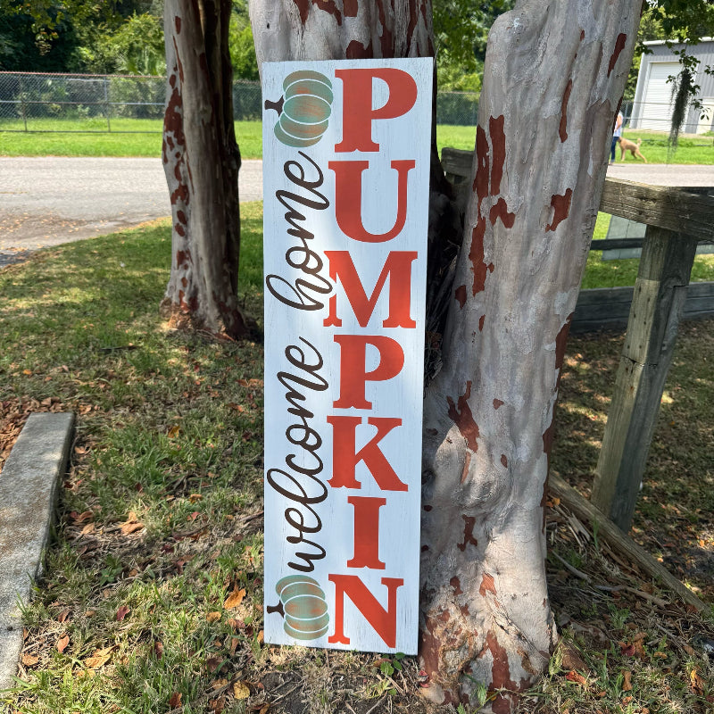 Wooden sign with 'Welcome Home Pumpkin' text and pumpkin graphics leaning against a tree.