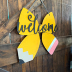 A decorative hanging sign shaped like a pencil with the word "welcome" in black script across the center, shown against a dark wood plank wall.