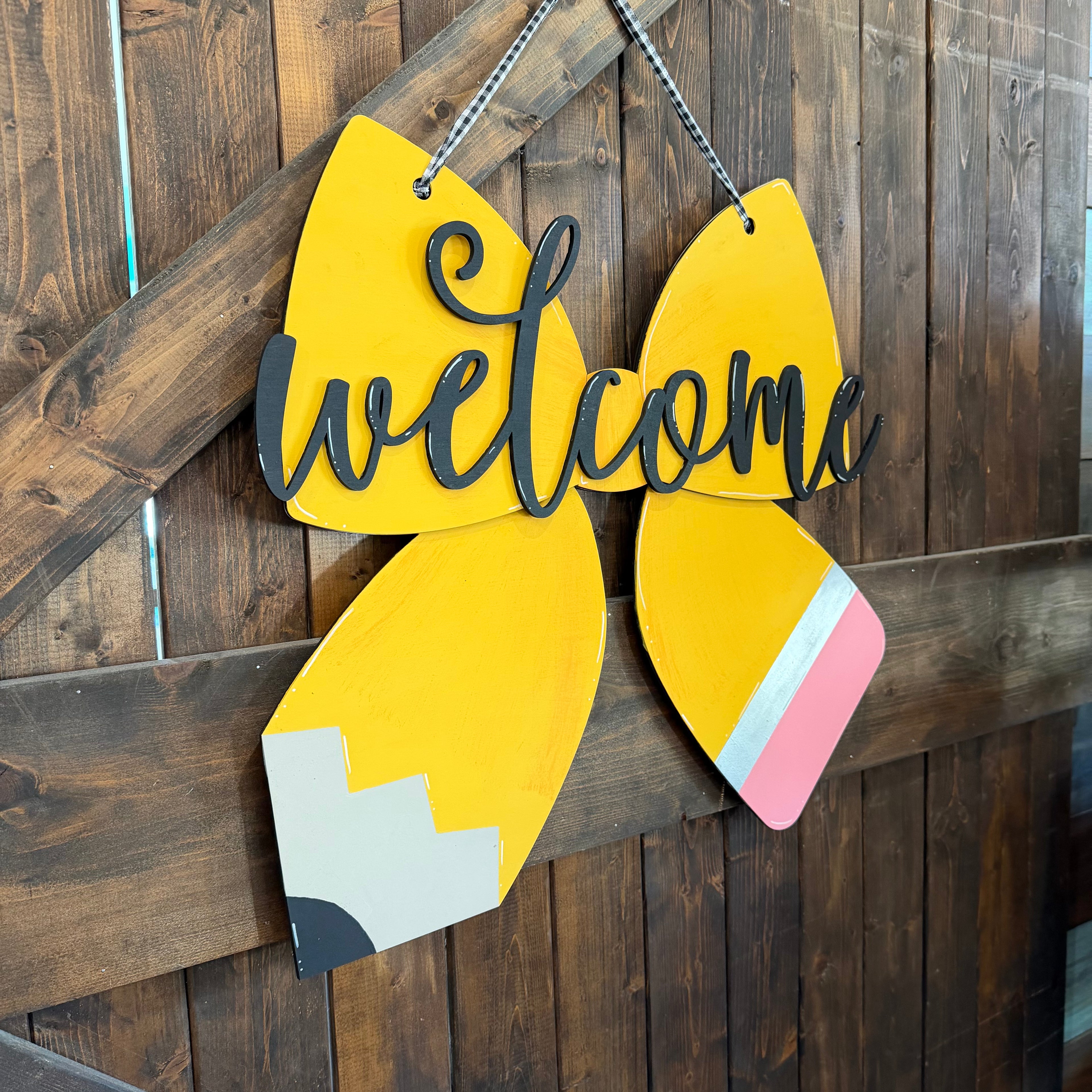 A decorative hanging sign shaped like a pencil with the word "welcome" in black script across the center, shown against a dark wood plank wall.