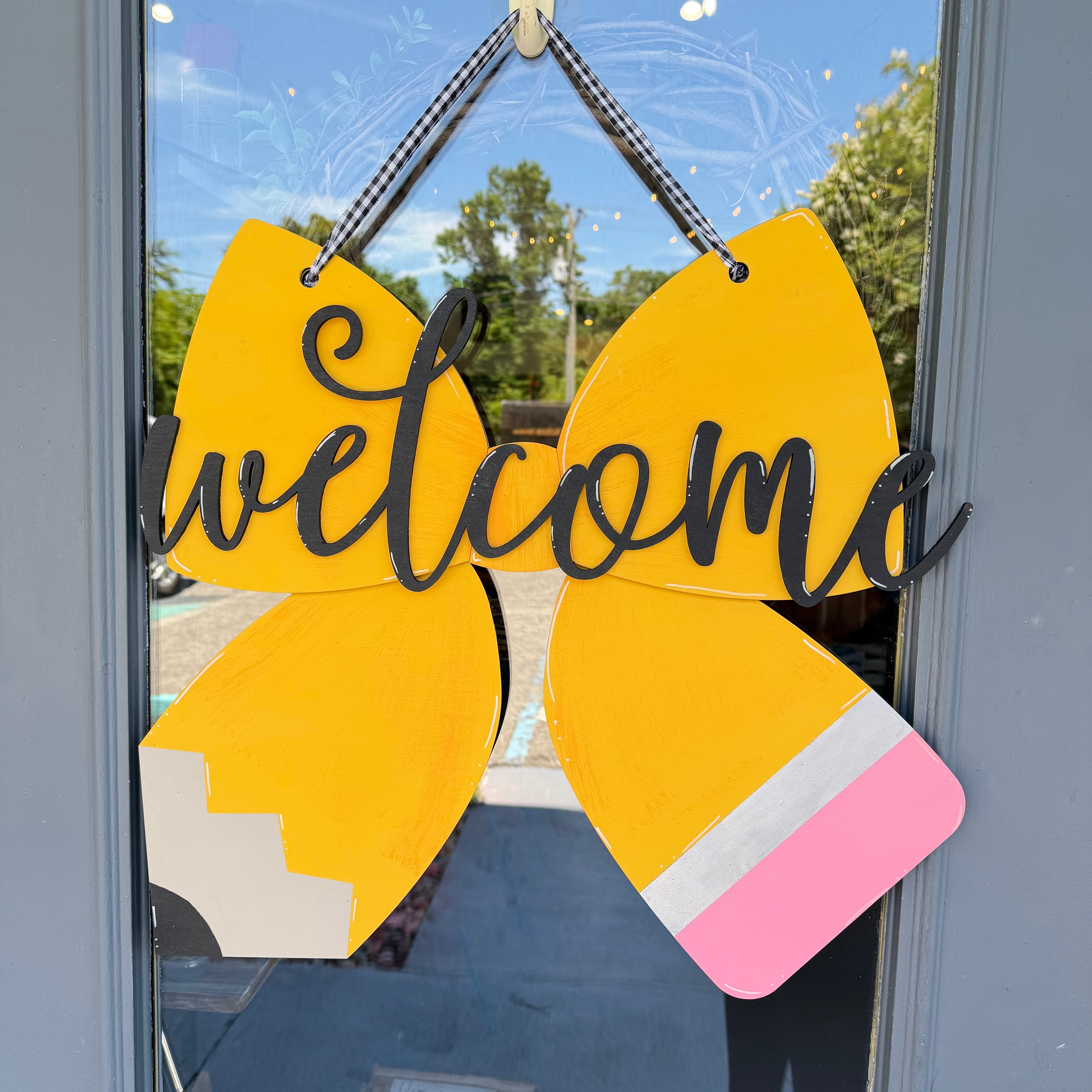 A decorative yellow bow-shaped hanging sign with a black script 'welcome' overlay, attached to a glass door. The bow is styled like a pencil with a cream section on one side, a pink eraser on the other, and a white center stripe.