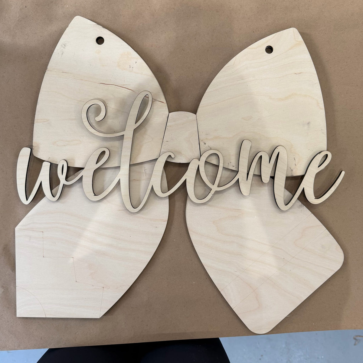 Unpainted wooden 'welcome' sign with bow-shaped panels and cursive script lettering, displayed on a brown surface.