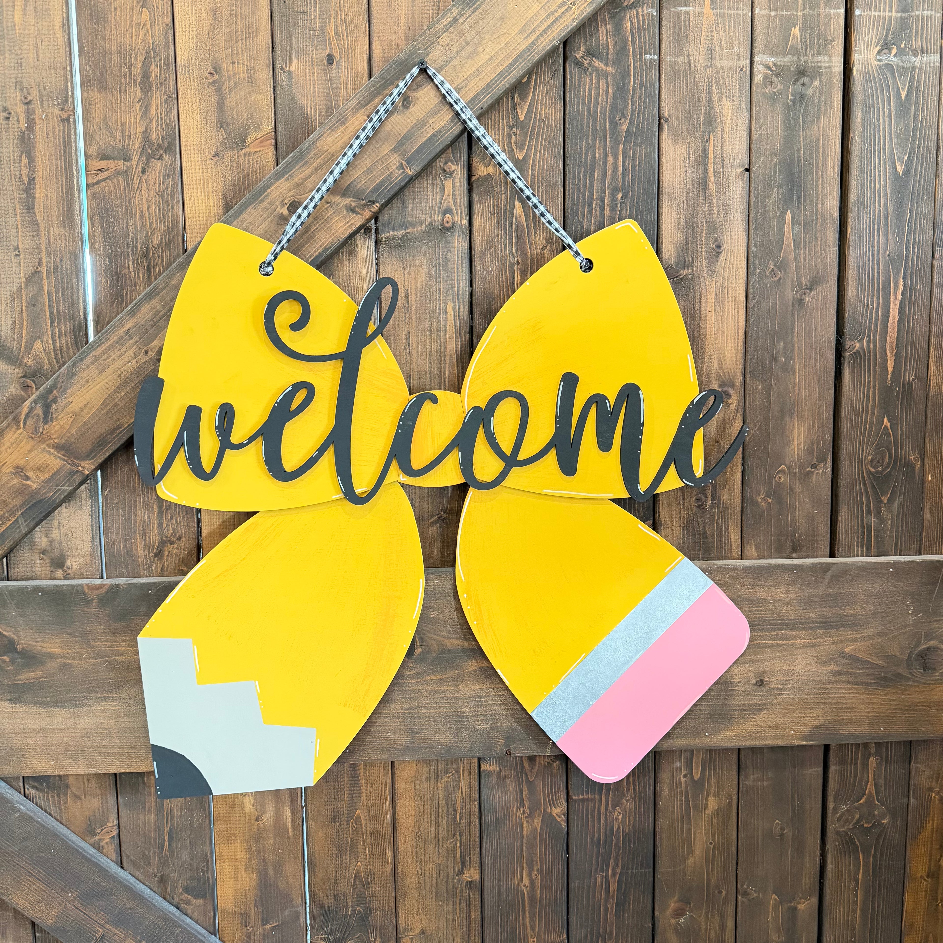 A decorative wooden hanging sign shaped like a yellow pencil with the word "welcome" in black script across the center, displayed against a rustic dark wood plank background.
