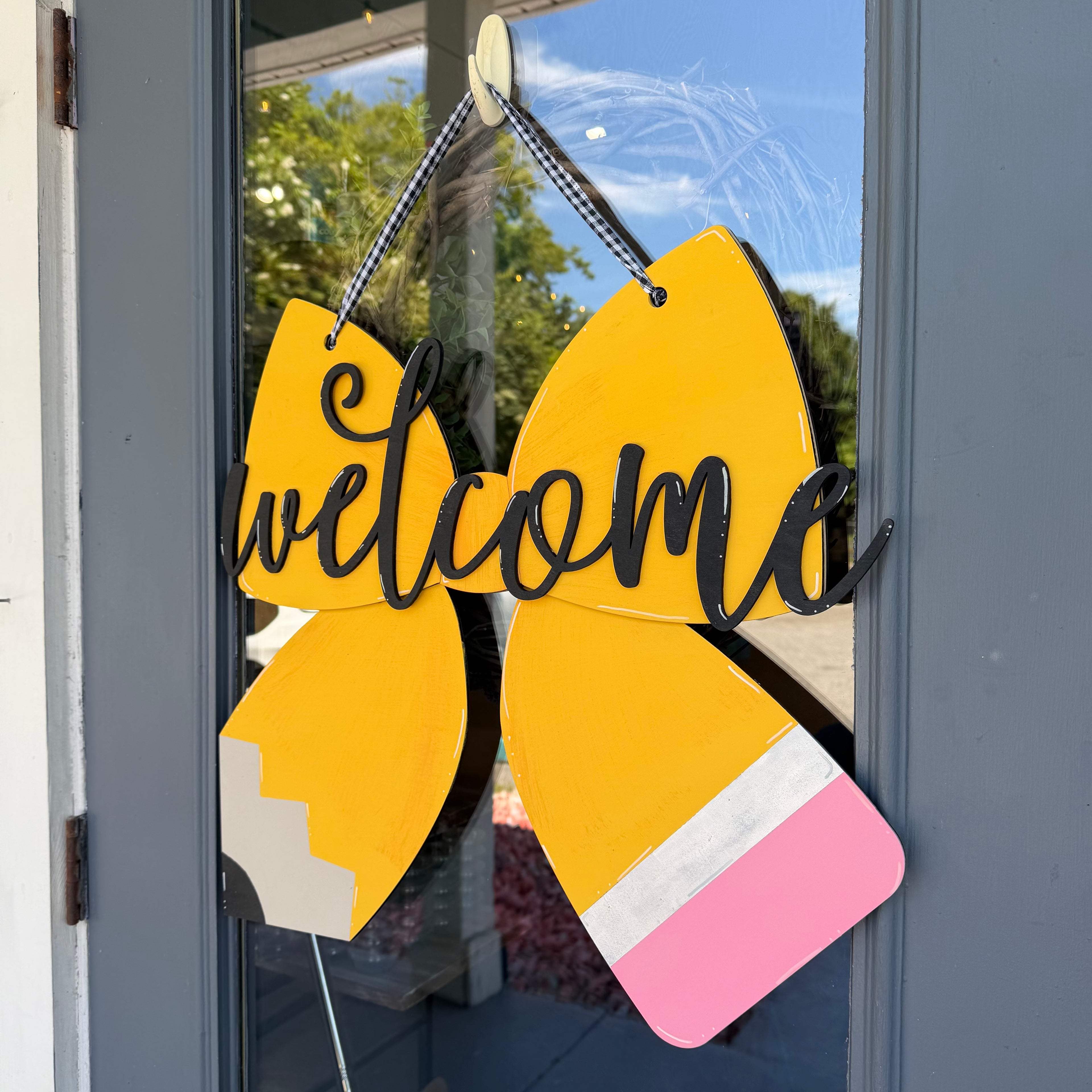 A decorative wooden sign shaped like a pencil with the word 'welcome' in black script across the center, hanging on a glass door. The sign is yellow with a pink eraser, a white center stripe, and a black-and-white checkered hanging ribbon.