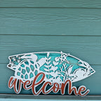 Decorative metal sign with fish design and 'welcome' text on a teal wooden background