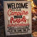 Wooden sign with text 'Welcome to our campfire where friends & marshmallows get toasted at the same time' in front of a campfire.